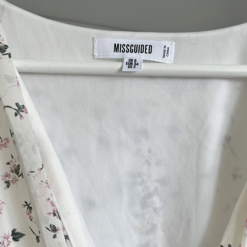 Säljer en vacker blommig klänning från Missguided i storlek XS. Klänningen har en omlott-design med v-ringning och långa ärmar. Den är vit med ett fint blommönster i rosa och grönt. Perfekt för vår och sommar!. Mekot.