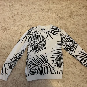 Vit tröja med svart palmbladsmönster från Your Turn - Snygg vit tröja med svart palmbladsmönster från Your Turn. Tröjan har långa ärmar och en bekväm passform. Perfekt för en avslappnad stil.