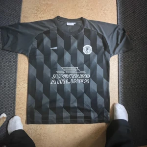 Svart och grå fotbollströja med mönster - Snygg svart och grå fotbollströja med diagonalt mönster och tryck från Junkyard Airlines. Tröjan har korta ärmar och ett stilrent tryck på framsidan. Perfekt för fotbollsfans som vill sticka ut på planen eller läktaren.