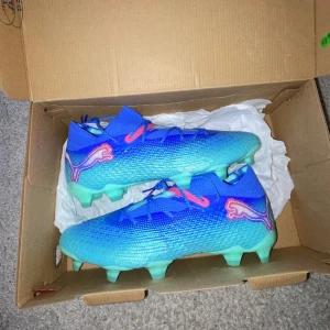 Blå och turkosa fotbollsskor från Puma - Snygga fotbollsskor från Puma ( Future elit ) i en cool blå och turkos färgkombination. Skorna har en strömlinjeformad design med snörning och en distinkt logotyp på sidan. Perfekta för att dominera på planen med stil. Direkt ifrån boxen, inte använda. 