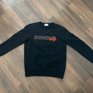 Svart sweatshirt från Dondup - Säljer en stilren svart sweatshirt från Dondup med tryck i vitt och orange på bröstet. Tröjan har långa ärmar och en rund halsringning. Perfekt för en avslappnad look.
