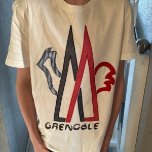 Moncler T-shirt - Säljer en snygg vit t-shirt från Moncler med ett stort tryck av 'Grenoble' i rött, blått och svart på framsidan. T-shirten har en klassisk rund hals och korta ärmar. Perfekt för en stilren look! Hör av er vid frågor! självklart äkta 