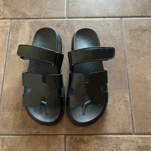 Svarta sandaler med justerbart spänne - Säljer dessa snygga svarta sandaler i minimalistisk design. De är i fint skick, endast använda ett fåtal gånger. Modellen har en stilren form med ett H-liknande snitt framtill och ett praktiskt kardborrespänne över foten för optimal passform. Perfekta för både vardag och semester! Står storlek 40 under men passar som 38,5.