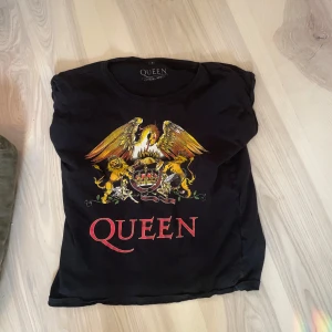 Svart Queen t-shirt - Säljer en svart t-shirt från Queen med ett coolt tryck av bandets emblem i gult och rött. Perfekt för alla Queen-fans som vill visa sin kärlek till bandet. T-shirten har korta ärmar och en normal passform.