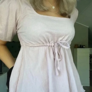 Ljusrosa sommarblus med knytband - Söt ljusrosa sommarblus med knytband i midjan och korta puffärmar. Den är ny utan prislapp och har endast provats, den säljs ej längre! Perfekt för en somrig look med en fyrkantig urringning. 