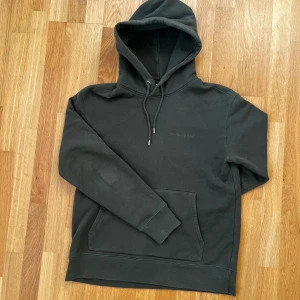 Mörkgrön hoodie från J.Lindeberg - Säljer en stilren mörkgrön hoodie från J.Lindeberg. Den har en klassisk design med justerbar huva och en praktisk magficka. Perfekt för en avslappnad look. Märkesloggan är diskret placerad på bröstet.