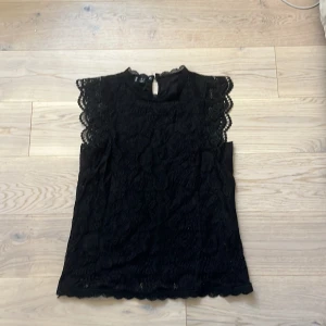 Svart spetsblus från Pieces - Säljer en elegant svart spetsblus från Pieces. Blusen har vackra spetsdetaljer och en knapp i nacken. Perfekt för att ge en stilren look.