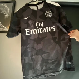 Svart PSG fotbollströja - Säljer en svart PSG fotbollströja från Nike med korta ärmar. Tröjan har ett kamouflagemönster och vit text med 'Fly Emirates' på framsidan. Baksidan har 'Neymar Jr' och nummer 10 i vitt och rött. Perfekt för fotbollsfans!