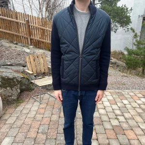 Barbour - Mörkblå quiltad hybrid jacka med dragkedja - Snygg mörkblå quiltad hybrid jacka med dragkedja framtill. Västen har ribbade muddar och en stilren design som passar perfekt för kyligare dagar. Nypris ligger runt 3000kr. Jackan är i ett mycket fint skick och storleken är medium.