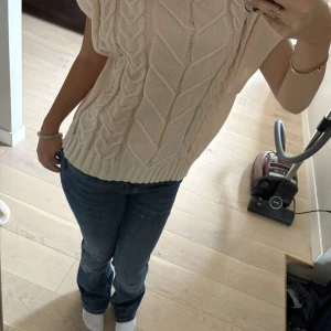 Beige stickad topp - Säljer en snygg beige stickad topp med korta ärmar och ett flätmönster framtill. Toppen har en ribbad kant vid ärmslut och nederkant, vilket ger en fin passform. Perfekt för en stilren look.