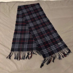 Rutig halsduk från Burberry - Snygg rutig halsduk från Burberry i svart, grått och rött. Tillverkad av 100% lammull, perfekt för att hålla dig varm under kyliga dagar. Klassisk design med fransar i ändarna.