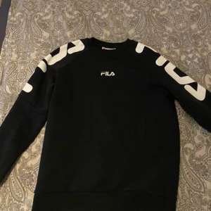 Svart sweatshirt från FILA - Snygg svart sweatshirt från FILA med vita detaljer på ärmarna och logga på bröstet. Perfekt för en avslappnad stil. Tröjan har långa ärmar och en bekväm passform.