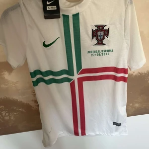 Vit Portugal fotbollströja från Nike - Säljer en vit Portugal fotbollströja från Nike med gröna och röda detaljer. Tröjan har korta ärmar och ett tryck med datumet 27/06/2012. Perfekt för fotbollsfans som vill visa sitt stöd för Portugal! 🇵🇹