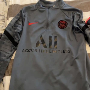Grå fotbollströja från Nike - Säljer en grå fotbollströja från Nike med PSG-logga och Accor Live Limitless-tryck. Tröjan har en halv dragkedja och långa ärmar med ett mönster på ärmarna. Perfekt för fotbollsfans!