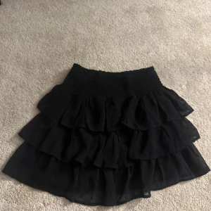 Svart volangkjol från Lindex - Snygg svart kortkjol från Lindex med flera lager av volanger. Perfekt för att ge en lekfull touch till din outfit. Kjolen har en elastisk midja för extra komfort.
