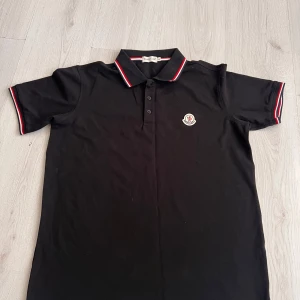 Svart pikétröja från Moncler - Snygg svart pikétröja från Moncler med korta ärmar och klassisk krage. Tröjan har röda och vita detaljer på kragen och ärmsluten samt en broderad logotyp på bröstet. Perfekt för en stilren look.