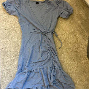 Blå kort klänning  - Säljer en blå klänning med korta ärmar och volangdetaljer. Klänningen har en knytning i midjan och en v-ringad. I storlek xxs.