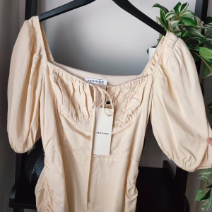 Beige klänning från Adoore i storlek 42 - Vacker ljusbeige sommarklänning från märket Adoore. Storlek 42. Denna klänningsmodell är snävare i midjepartiet. Adoore rekommenderar att du går upp 1 storlek. Det vill säga att du med storlek 40 borde kunna ha denna. Sen är alla olika men det är vad Adoore rekommenderar. Helt ny med lappar kvar. Har en dragsko på ena sidan så du kan välja längd på klänningen efter eget tycke. Knyte vid bysten. Även resår vi ärmarna. Dold dragkedja vid sidan. Fin korsett-liknande överdel.