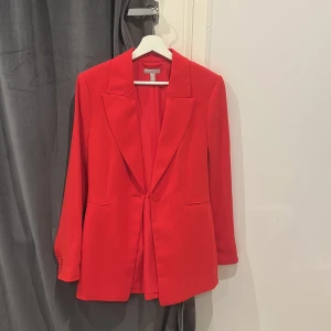 Röd kavaj från H&M - Snygg röd kavaj från H&M i storlek 38. Kavajen har en klassisk design med långa ärmar och knappar vid ärmsluten. Perfekt för att ge en färgklick till din outfit. Den har en enkelknäppning framtill och är tillverkad i ett mjukt material. Använd endast vid 1 tillfälle. 