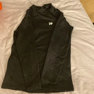 Svart långärmad träningströja - Säljer en svart långärmad träningströja med ett diskret mönster på ärmarna. Perfekt för träning och andra sportaktiviteter. Tröjan har en enkel design med en liten logotyp på bröstet.