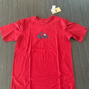Röd t-shirt från Quiksilver - Säljer en röd t-shirt från Quiksilver med deras ikoniska logga tryckt på bröstet. T-shirten är kortärmad och gjord i mjuk bomull, perfekt för en avslappnad stil. Passar bra till jeans eller shorts för en cool look. Tröjan är aldrig använd och i perfekt skick! Pris kan diskuteras. Storlek XL men passar mer som S-M