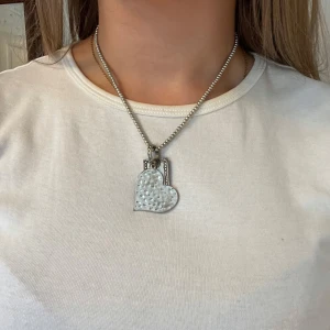 Silverfärgat halsband med hjärta och tag - Unikt halsband i silverfärg med kulkedja och två hängen: ett hamrat hjärta och en rektangulär tag med texten 'joy'. Perfekt statement-piece för dig som gillar personliga smycken med cool vibe. Ganska väl använt vilket syns då det är lite slitet.