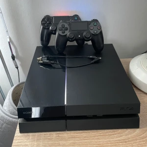 Ps4 - Bilderna visar en PlayStation 4-konsol med två svarta DualShock 4-kontroller och en laddningskabel. Perfekt för gamingkvällar med vänner! Konsolen och kontrollerna ser ut att vara i gott skick och redo för nya äventyr. Ett måste för dig som älskar spel och vill ha en klassisk spelupplevelse.
