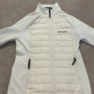 Vit  jacka/fleece Peak Performance  - Snygg vit jacka från Peak Performance i storlek L. Jackan har quiltade paneler fram och bak, dragkedja hela vägen och två fickor med dragkedja. Perfekt för kyliga dagar och har en sportig vibe med hög krage och stilren design.