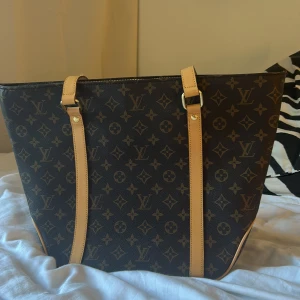 Louis Vuitton Monogram axelväska - Säljer en klassisk Louis Vuitton axelväska i brunt monogrammönster med ljusa skinnremmar och guldfärgade metalldetaljer. Väskan har rymlig form och ikoniska LV-loggor över hela ytan. Perfekt för dig som vill ha en statementväska med lyxig känsla.