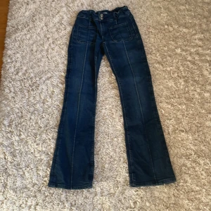 Blå bootcut jeans från Gina Tricot - Snygga blå jeans från Gina Tricot i storlek 158. Modellen har bootcut-ben, hög midja och stora framfickor. Jeansen har markerade sömmar framtill och stängs med två knappar. Perfekta för en trendig och avslappnad look.