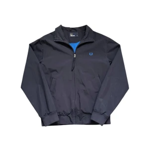 vindjacka från Fred Perry - Klassisk vindjacka från Fred Perry med blått broderat logomärke på bröstet. Jackan har dragkedja framtill, två sidofickor och ribbade muddar vid ärmslut och midja. Perfekt för säsongens alla dagar.