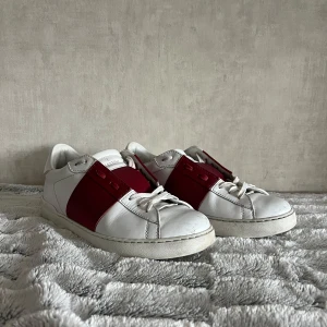 Valentino Open sneakers vit/röd - Säljer ett par Valentino Open sneakers i vitt skinn med bred röd läderdetalj över snörningen. Skorna har platt sula och ikoniska nitar bak på hälen. Snygg och stilren design som sticker ut med den röda färgblocken. Perfekta för dig som gillar lyxiga sneakers.