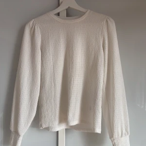 Vit långärmad topp från Vero Moda - Superfin vit långärmad topp från Vero Moda med lätt struktur i tyget. Klassisk rund halsringning och diskret puff vid axlarna ger en trendig touch. Perfekt att styla med jeans eller kjol för en clean look.