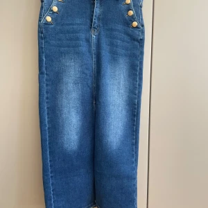 Blå jeanskjol med knappar 36 - Snygg blå lång jeanskjol  med dekorativa guldknappar framtill och slits. Klassisk femficksmodell och hög midja. Materialmix av bomull och polyester med lite stretch för skön passform.