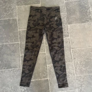 Gymshark tights  - Säljer ett par helt nya och oanvända Gymshark-tights i storlek S. Dessa tights har ett snyggt kamouflagemönster. Perfekta för gymmet, löprundan eller bara som bekväma vardagsbyxor. Gymshark-loggan finns synlig på vänster lår.
