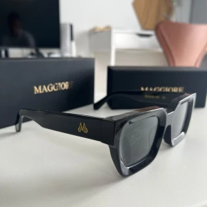 Svarta solglasögon från Maggiore - Stilrena svarta solglasögon från Maggiore med rektangulära bågar och mörka glas. Snygg gulddetalj med logga på skalmen och robust plastmaterial. Perfekta för dig som vill ha en modern och edgy look.
