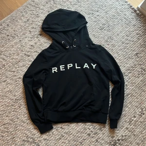 Svart hoodie från Replay med tryck - Svart hoodie från Replay med stor vit logga framtill. Klassisk modell med huva och snörning, ribbade muddar och lång ärm. Perfekt för en chill och stilren look. Mjukt material som känns skönt mot huden.