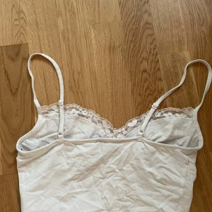 Vit spetslinnetopp med rosett - Supersöt vit linnetopp med smala axelband och spetsdetaljer längs bysten. Toppen har en liten rosett framtill och blommig spets som ger en romantisk vibe. Perfekt till jeans eller kjol för en söt look.
