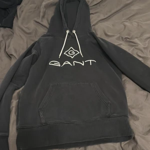 Svart hoodie från GANT med logga - Svart hoodie från GANT med stor vit logga och emblem på bröstet. Tröjan har huva med snörning och en klassisk känguruficka framtill. Ribbad mudd vid ärmslut och nederkant. väldigt bra skick