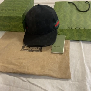 Svart keps från Gucci med GG-mönster - Svart keps från Gucci med diskret GG-monogram över hela ytan och klassiska gröna och röda ränder på sidan. Justerbar rem i svart skinn baktill. Kepsen är tillverkad i bomull och har en snygg, sportig look med exklusiv känsla.
