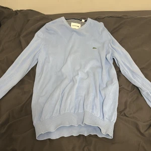 Ljusblå stickad tröja från Lacoste - Snygg ljusblå stickad tröja från Lacoste med klassisk krokodillogga på bröstet. Tröjan har ribbade muddar vid ärmslut och nederkant samt rund hals. Perfekt för dig som gillar stilrena och softa plagg med premiumkänsla. mycket bra skick