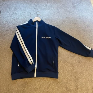  Palm Angels kofta - Säljer en snygg marinblå ziptröja från Palm Angels med vita ränder längs ärmarna och vit dragkedja. Tröjan har hög krage, två fickor med dragkedja och logga på bröstet. Perfekt för en sportig och trendig look.