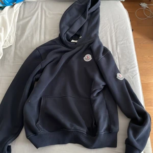 Svart hoodie från Moncler med logga - Snygg blå hoodie från Moncler med klassisk logga på bröstet och ärmen. Tröjan har huva, känguruficka och ribbade muddar. Perfekt för dig som gillar stilrena och exklusiva plagg med streetkänsla.