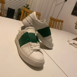 Gröna Valentino opens - Snygga Valentino Garavani opens sneakers i vitt skinn med bred grön rem över vristen och coola nitar på hälen. Klassisk rund tå, vita snören och platt sula. Perfekta för dig som vill sticka ut med exklusiva detaljer och stilren design.