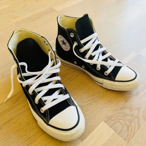 Svarta Converse Chuck Taylor All Star - Klassiska svarta Converse Chuck Taylor All Star high-tops med vit sula och tå, vita snören och tydliga kontrastsömmar. Ikonisk logga på sidan och metallöljetter. Ovandel i canvas och platt gummisula. Perfekta för en cool och avslappnad stil.