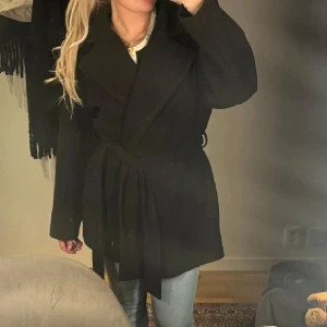 Svart omlottkappa med bälte - Stilren svart kappa med bred krage och knytbälte i midjan. Jackan har en omlottmodell och raka, långa ärmar. Perfekt för dig som gillar en clean och klassisk look. Materialet känns mjukt och ger en snygg siluett.