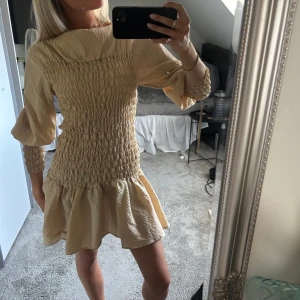 Beige kort klänning med volang - Populär smockad beige kort klänning och volangig kjol. Långa puffärmar med smock vid handlederna och hög hals. Materialet är lätt och har en krinklad struktur som ger en cool och trendig vibe.