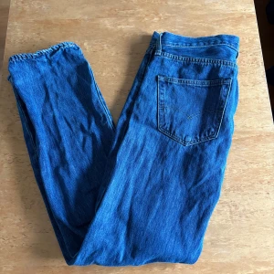 Levi's 501 blå jeans W32 L32 - Klassiska Levi's 501 jeans i blå denim. Rak passform med fem fickor och knappgylf. Snyggt mellanblå färg och ikonisk läderpatch bak i midjan. Perfekta för dig som gillar tidlös stil och vill ha ett par riktigt schyssta jeans.
