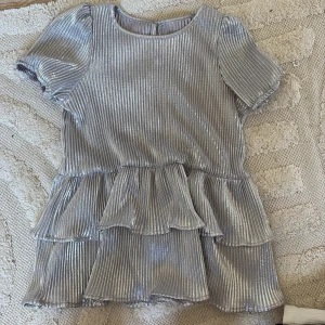 Glittrig silver blus med volanger - Supersöt blus som egentligen är en bäbiskläning men passar även som en fin blus, tyget är stretchigt och därför passar perfekt som en blus jag har storlek 146/152 och passar perfekt på mig så rekommenderar att ha storlek 126/152.👍 blusen har flera lager volanger som ger en riktigt festlig känsla Perfekt för dig som vill sticka ut och glänsa lite extra!🤗