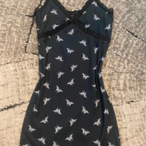 Svart kort klänning med fjärilar H&M, S  - Supersöt svart kort klänning från H&M Divided med vitt fjärilsmönster. Klänningen har spetsdetaljer vid halsringningen och en liten rosett framtill. Den är ärmlös och har en slim passform som framhäver figuren. Perfekt för dig som gillar unika prints och detaljer.